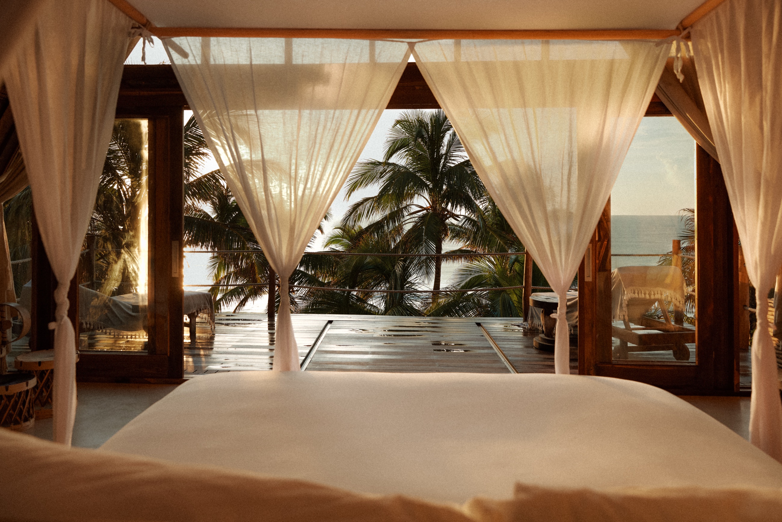 View through a sheer curtain-draped canopy bed to palm trees and the ocean at sunset, Namron Hotels, Tulum